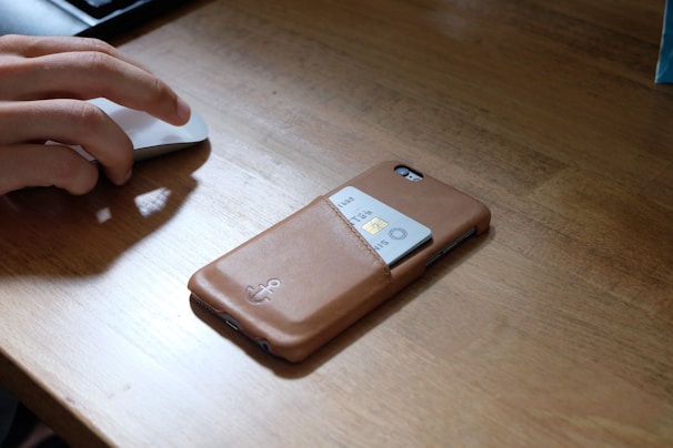 A person's hand is using a computer mouse on a wooden desk. Next to the hand, there is a smartphone with a brown leather case. The case has a slot holding a credit card partially visible.