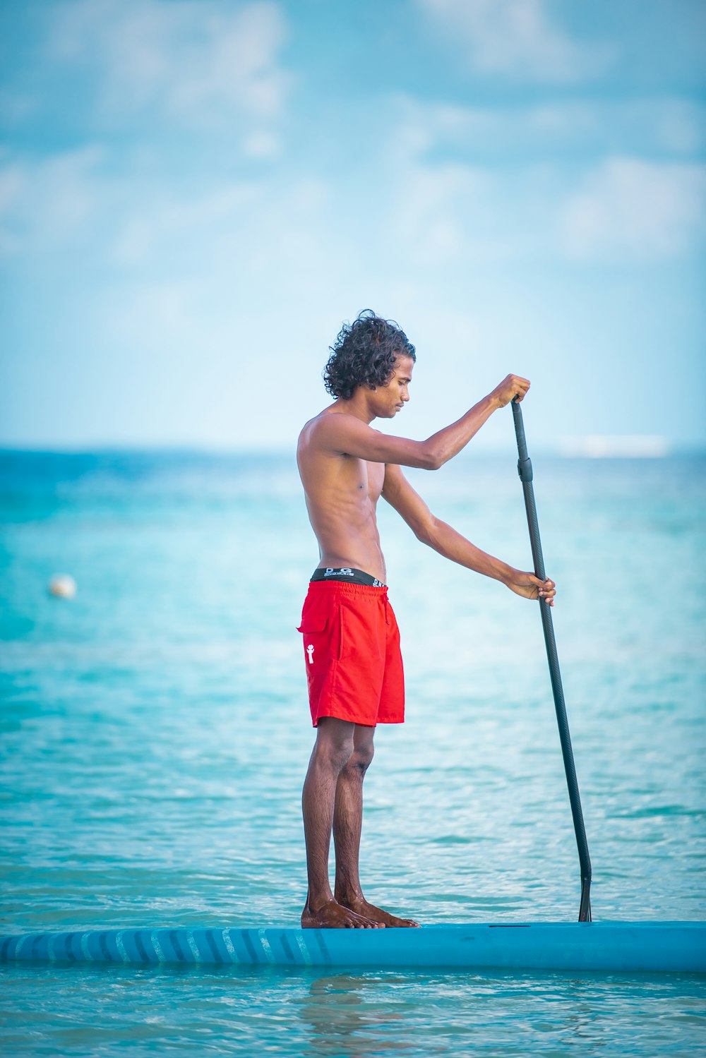topless boy in red shorts holding black and white stick standing on body of water during