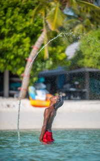 A person stands waist-deep in clear blue water, wearing red shorts. They are tilting their head back, allowing a stream of water to arc from their mouth in a playful manner. Lush green palm trees and beach huts are visible in the background, indicating a tropical setting.