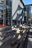 A modern architectural structure includes a large, textured facade casting intricate shadows. In the foreground, there are tiered concrete benches made of stone or concrete. The building's extensive use of glass creates an open and airy aesthetic, while the light and shadows add depth.