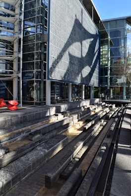 A modern architectural structure includes a large, textured facade casting intricate shadows. In the foreground, there are tiered concrete benches made of stone or concrete. The building's extensive use of glass creates an open and airy aesthetic, while the light and shadows add depth.