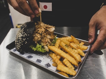 A hand is holding a small flag with the Japanese symbol, placed into a unique burger with a black sesame seed bun, lettuce, and a generous amount of toppings. The dish is served on a tray with crinkle-cut fries beside it.