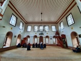 An interior of a mosque with high ceilings and patterned windows. The room features several people sitting on a carpeted floor facing a wooden pulpit. The architecture includes decorative elements such as chandeliers and arched windows.
