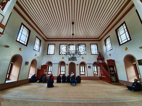 An interior of a mosque with high ceilings and patterned windows. The room features several people sitting on a carpeted floor facing a wooden pulpit. The architecture includes decorative elements such as chandeliers and arched windows.