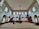 An interior of a mosque with high ceilings and patterned windows. The room features several people sitting on a carpeted floor facing a wooden pulpit. The architecture includes decorative elements such as chandeliers and arched windows.