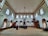 An interior of a mosque with high ceilings and patterned windows. The room features several people sitting on a carpeted floor facing a wooden pulpit. The architecture includes decorative elements such as chandeliers and arched windows.
