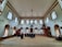 An interior of a mosque with high ceilings and patterned windows. The room features several people sitting on a carpeted floor facing a wooden pulpit. The architecture includes decorative elements such as chandeliers and arched windows.