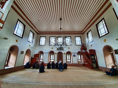 An interior of a mosque with high ceilings and patterned windows. The room features several people sitting on a carpeted floor facing a wooden pulpit. The architecture includes decorative elements such as chandeliers and arched windows.