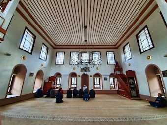 An interior of a mosque with high ceilings and patterned windows. The room features several people sitting on a carpeted floor facing a wooden pulpit. The architecture includes decorative elements such as chandeliers and arched windows.