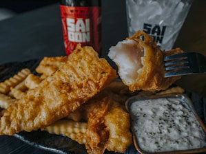 A plate of crispy, golden-brown fried fish and crinkle-cut fries is accompanied by a small dish of tartar sauce. A piece of fish is held up by a fork, showcasing its flaky texture. In the background, there is a bottle and a glass with ice, featuring the logo 'SAI GON'.