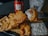 A plate of crispy, golden-brown fried fish and crinkle-cut fries is accompanied by a small dish of tartar sauce. A piece of fish is held up by a fork, showcasing its flaky texture. In the background, there is a bottle and a glass with ice, featuring the logo 'SAI GON'.