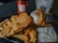 A plate of crispy, golden-brown fried fish and crinkle-cut fries is accompanied by a small dish of tartar sauce. A piece of fish is held up by a fork, showcasing its flaky texture. In the background, there is a bottle and a glass with ice, featuring the logo 'SAI GON'.