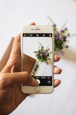 A hand is holding a smartphone, capturing an image of a notebook, a pen, and a small bouquet of purple flowers on a white surface. The smartphone screen shows the image being taken with a visible camera interface.