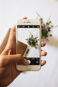 A hand is holding a smartphone, capturing an image of a notebook, a pen, and a small bouquet of purple flowers on a white surface. The smartphone screen shows the image being taken with a visible camera interface.