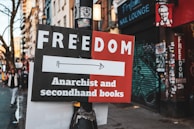 A street sign attached to a pole points towards a direction, advertising a place that offers 'Anarchist and secondhand books.' The sign is divided into two colors: black and red. In the background, there are buildings with graffiti on the shutters and walls, including a nail lounge. There is a faded sticker on the pole and a few pedestrians walking on the sidewalk.