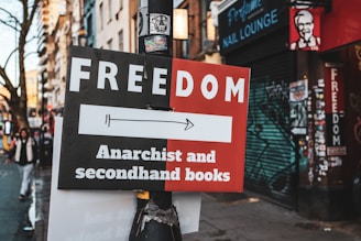 A street sign attached to a pole points towards a direction, advertising a place that offers 'Anarchist and secondhand books.' The sign is divided into two colors: black and red. In the background, there are buildings with graffiti on the shutters and walls, including a nail lounge. There is a faded sticker on the pole and a few pedestrians walking on the sidewalk.