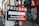 A street sign attached to a pole points towards a direction, advertising a place that offers 'Anarchist and secondhand books.' The sign is divided into two colors: black and red. In the background, there are buildings with graffiti on the shutters and walls, including a nail lounge. There is a faded sticker on the pole and a few pedestrians walking on the sidewalk.