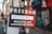 A street sign attached to a pole points towards a direction, advertising a place that offers 'Anarchist and secondhand books.' The sign is divided into two colors: black and red. In the background, there are buildings with graffiti on the shutters and walls, including a nail lounge. There is a faded sticker on the pole and a few pedestrians walking on the sidewalk.