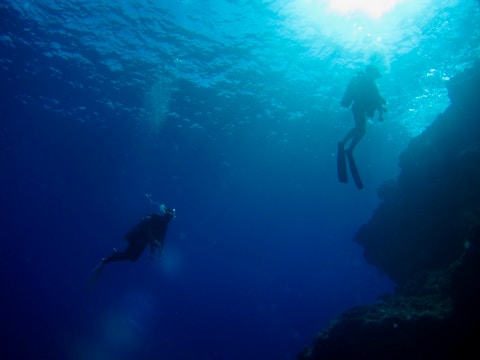 Two divers are exploring underwater, surrounded by deep blue water and illuminated by sunlight filtering from above. Their silhouettes are visible as they swim near a rocky underwater structure.