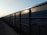 A row of freight train cars extends into the distance along a railway track. The cars are large, rectangular, and made of metal, with a blue-gray color. The lighting suggests it is either early morning or late afternoon, casting shadows along the textured surface of the cars.