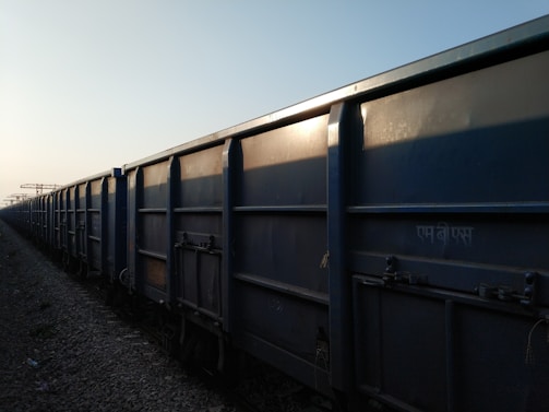 A row of freight train cars extends into the distance along a railway track. The cars are large, rectangular, and made of metal, with a blue-gray color. The lighting suggests it is either early morning or late afternoon, casting shadows along the textured surface of the cars.