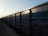 A row of freight train cars extends into the distance along a railway track. The cars are large, rectangular, and made of metal, with a blue-gray color. The lighting suggests it is either early morning or late afternoon, casting shadows along the textured surface of the cars.