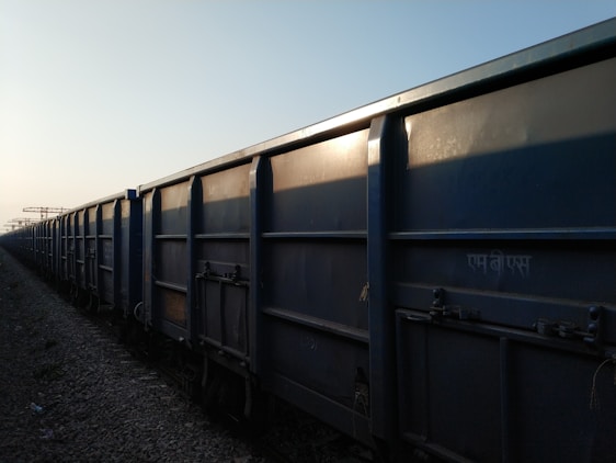 A row of freight train cars extends into the distance along a railway track. The cars are large, rectangular, and made of metal, with a blue-gray color. The lighting suggests it is either early morning or late afternoon, casting shadows along the textured surface of the cars.