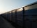A row of freight train cars extends into the distance along a railway track. The cars are large, rectangular, and made of metal, with a blue-gray color. The lighting suggests it is either early morning or late afternoon, casting shadows along the textured surface of the cars.