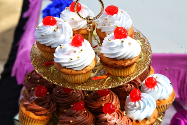 A tiered stand holding various cupcakes topped with swirls of white and chocolate frosting. Each cupcake is garnished with a red cherry and small gold decorative beads. The stand is ornate, made of gold, and positioned on a colorful table with purple and black elements in the background.