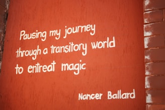 A brick wall painted red with the quote 'Pausing my journey through a transitory world to entreat magic' written in white letters. The signature 'Nancer Ballard' is at the bottom right.