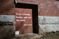 A section of a brick and stone wall with a wooden door painted in light brown. On the door, there's a handwritten quote in white: 'The gate to knowledge closed by a paper cover New realities Joshua Lozano.' The wall has a red brick pattern on the upper part and rough gray stone at the bottom.