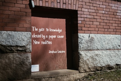 A section of a brick and stone wall with a wooden door painted in light brown. On the door, there's a handwritten quote in white: 'The gate to knowledge closed by a paper cover New realities Joshua Lozano.' The wall has a red brick pattern on the upper part and rough gray stone at the bottom.