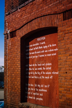 A brick wall with an arched section containing a wooden panel. The panel features text, including the title 'PERCEPTION'. The text appears to be a narrative or poem that reflects on natural phenomena and perception.