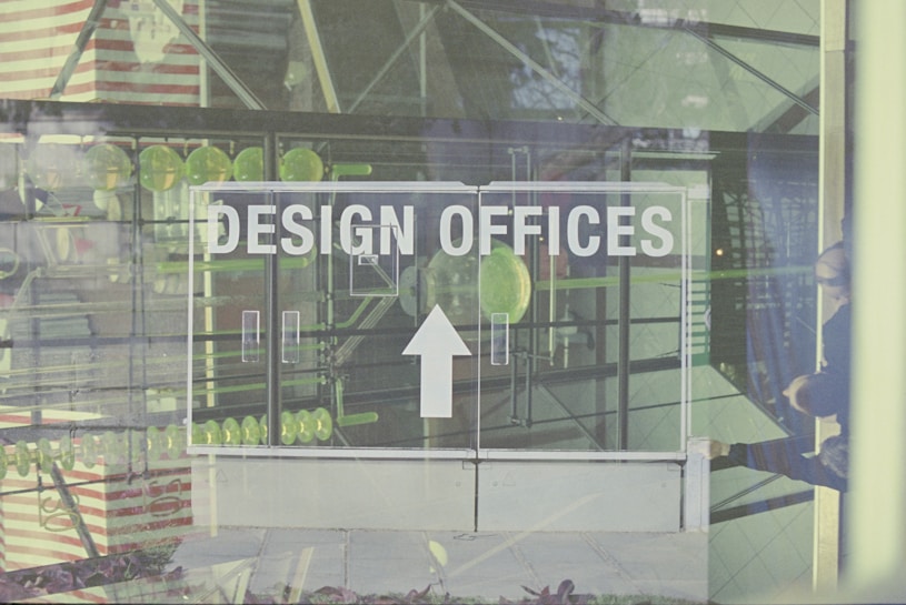 A glass door with the words 'Design Offices' printed on it is prominent in the foreground. Reflections on the glass reveal geometric structures and patterns, as well as hints of interior decor like lines of green circular objects. The faint outline of a person can be seen on the right side of the image, suggesting movement or activity behind the door.