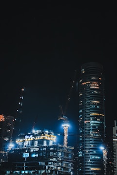 Nighttime cityscape showing modern buildings under construction with illuminated cranes.