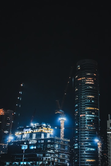 A striking nighttime view of a skyscraper under construction, illuminated by bright work lights against a deep blue sky.