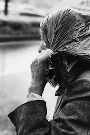 A person holding a plastic bag over their head, seemingly to shield from rain. The image is in black and white, focusing on the person's hand and the texture of the plastic bag and clothing. The setting appears to be outdoors, likely on a rainy day.