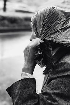 A person holding a plastic bag over their head, seemingly to shield from rain. The image is in black and white, focusing on the person's hand and the texture of the plastic bag and clothing. The setting appears to be outdoors, likely on a rainy day.