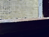 A close-up view of a wire mesh screen with a slightly blurred background. The mesh is dark and contrasts against the lighter background, which appears to be an out-of-focus exterior or natural light source. The image captures the grid-like pattern and the texture of the mesh.