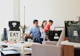 A young couple sits on a sofa with a toddler who is reaching into a black storage box labeled 'Hive Boxx'. The room has a cozy atmosphere with a well-lit background featuring a potted plant and a piece of framed art with the word 'EAT'. A white coffee table in the foreground displays various colorful toys and a mug.