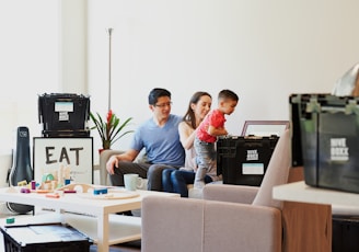 A young couple sits on a sofa with a toddler who is reaching into a black storage box labeled 'Hive Boxx'. The room has a cozy atmosphere with a well-lit background featuring a potted plant and a piece of framed art with the word 'EAT'. A white coffee table in the foreground displays various colorful toys and a mug.