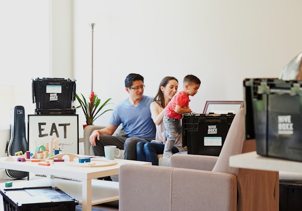 A young couple sits on a sofa with a toddler who is reaching into a black storage box labeled 'Hive Boxx'. The room has a cozy atmosphere with a well-lit background featuring a potted plant and a piece of framed art with the word 'EAT'. A white coffee table in the foreground displays various colorful toys and a mug.