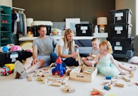 A family of four sits on a carpeted floor in a cozy room, surrounded by various toys and wooden building blocks. Parents are sitting with their two young children, all engaged in playing with the toys. The background features packed boxes labeled 'HIVE BO XX' and a shelf with clothes and other household items.