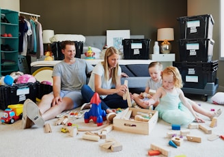 A family of four sits on a carpeted floor in a cozy room, surrounded by various toys and wooden building blocks. Parents are sitting with their two young children, all engaged in playing with the toys. The background features packed boxes labeled 'HIVE BO XX' and a shelf with clothes and other household items.