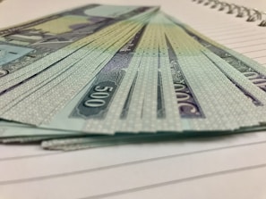 A stack of currency notes with a denomination of 500 is fanned out on a lined notebook. The design on the notes includes intricate patterns and a script-like font. The edges of the notebook pages are visible beneath the money.