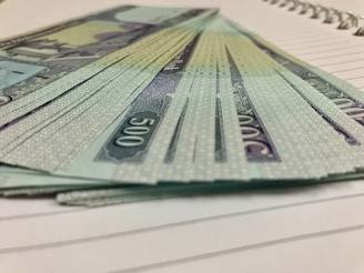 A stack of currency notes with a denomination of 500 is fanned out on a lined notebook. The design on the notes includes intricate patterns and a script-like font. The edges of the notebook pages are visible beneath the money.