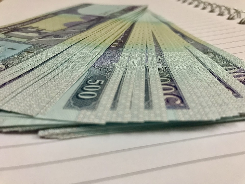 A stack of currency notes with a denomination of 500 is fanned out on a lined notebook. The design on the notes includes intricate patterns and a script-like font. The edges of the notebook pages are visible beneath the money.