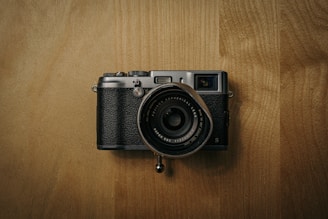 A vintage-style camera with a black textured body and metallic accents rests on a wooden surface. The lens, positioned centrally, is large and detailed, featuring clear labeling around its edges. The lighting creates a muted, warm ambiance.