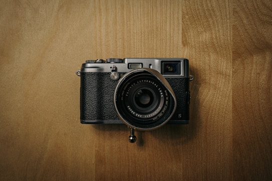 A vintage-style camera with a black textured body and metallic accents rests on a wooden surface. The lens, positioned centrally, is large and detailed, featuring clear labeling around its edges. The lighting creates a muted, warm ambiance.