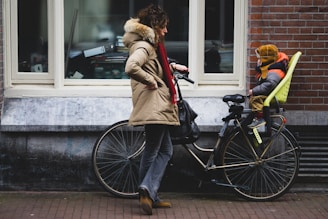 A person dressed in a winter coat stands beside a bicycle, engaging with a child who is sitting in a child seat mounted on the bike. The scene takes place on a city street with a brick wall and windows in the background.